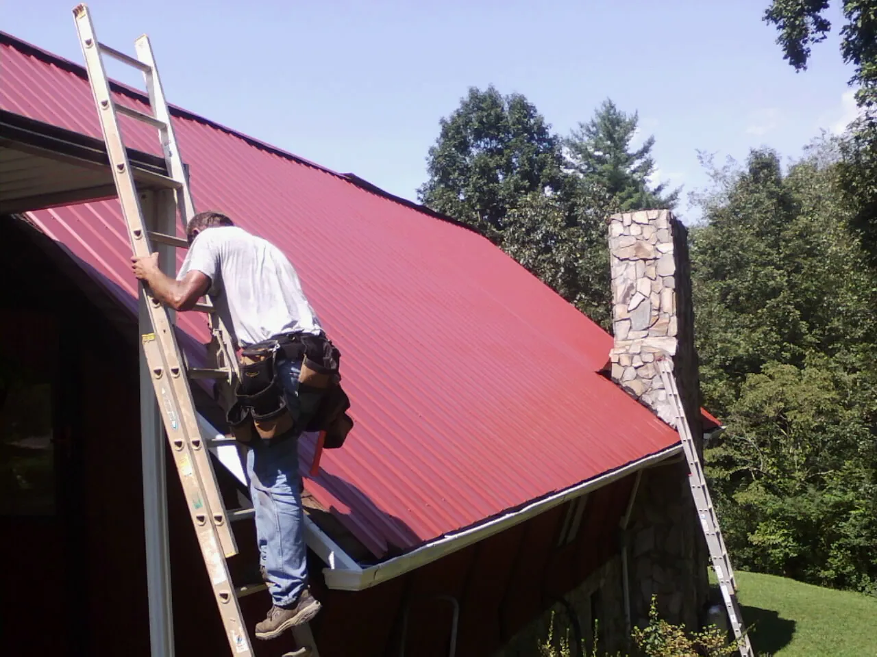 Roofer on a ladder beside a red metal roof and stone chimney — Soffit & Fascia Repair by Roofing Brothers in Colchester